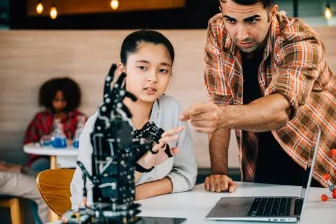 A student and instructor examine a robotic arm beside a laptop during a hands-on technology lesson, illustrating AI courses for the future.