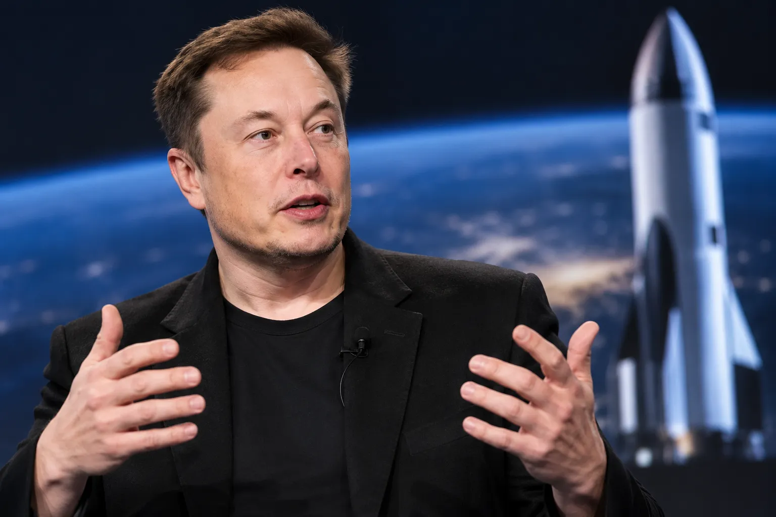 Elon Musk speaking with a rocket in the background